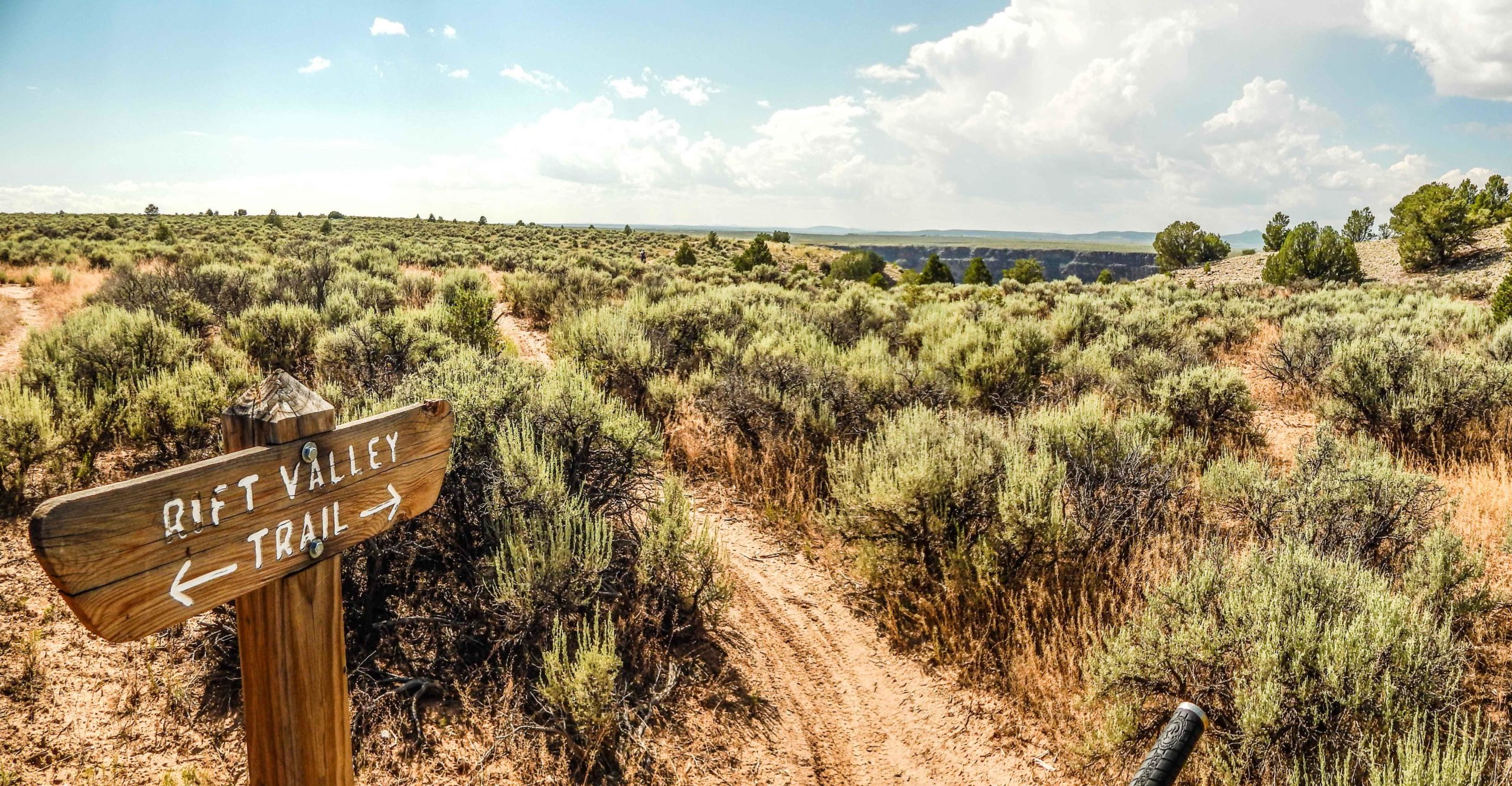 A wooden signpost for the "Rift Valley Trail" pointing left and right, set against a scenic view of a vast, grassy landscape with shrubs and distant hills under a partly cloudy sky. Taos Valley Overlook mountain bike trail.