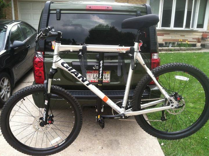 Giant Yukon: A silver mountain bike mounted on the back of a vehicle with a bike rack. The bike has large tires and front suspension, featuring the brand name "Giant" prominently displayed on the frame. In the background, a green SUV is visible with a partially visible license plate and a grassy area. The setting appears to be a driveway.
