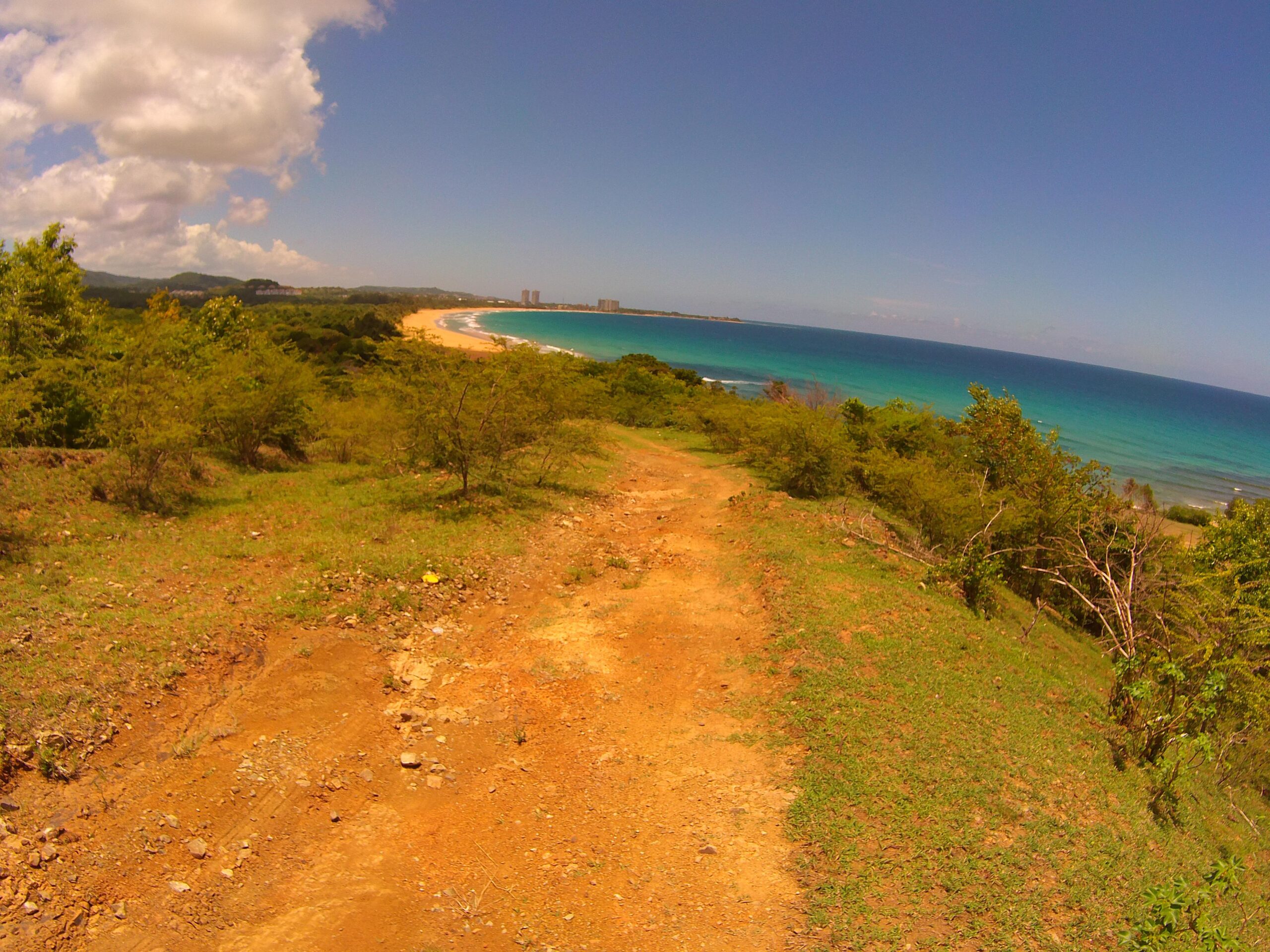 A scenic view from a hillside overlooking a sandy beach and turquoise ocean, with a dirt path leading down through green vegetation under a partly cloudy sky. Corredor Ecologico mountain bike trail.