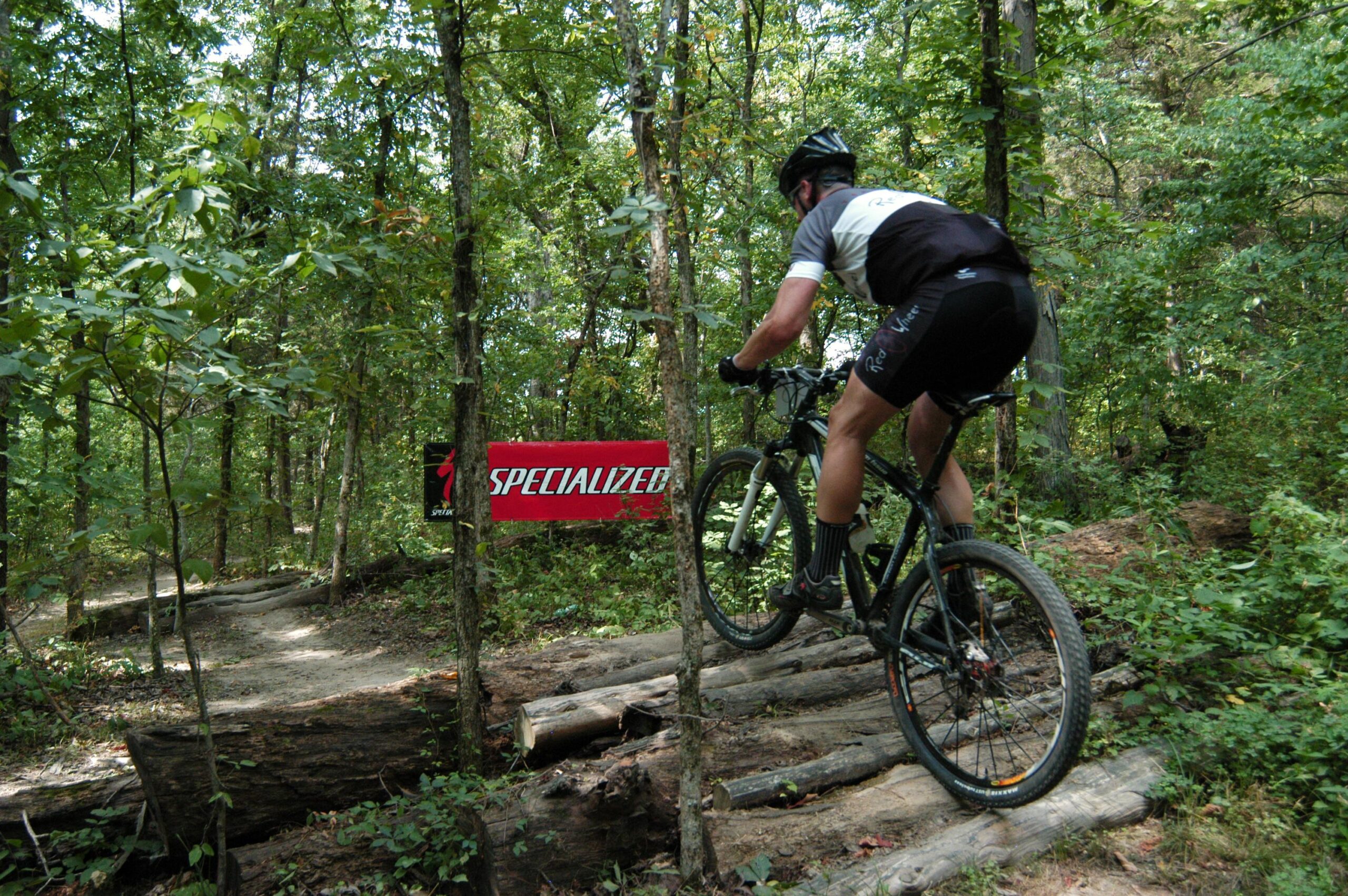 A mountain biker navigating over a series of logs on a forest trail, with a "Specialized" banner visible in the background. The scene is surrounded by lush green trees, showcasing a natural outdoor environment. Binder Lake mountain bike trail.