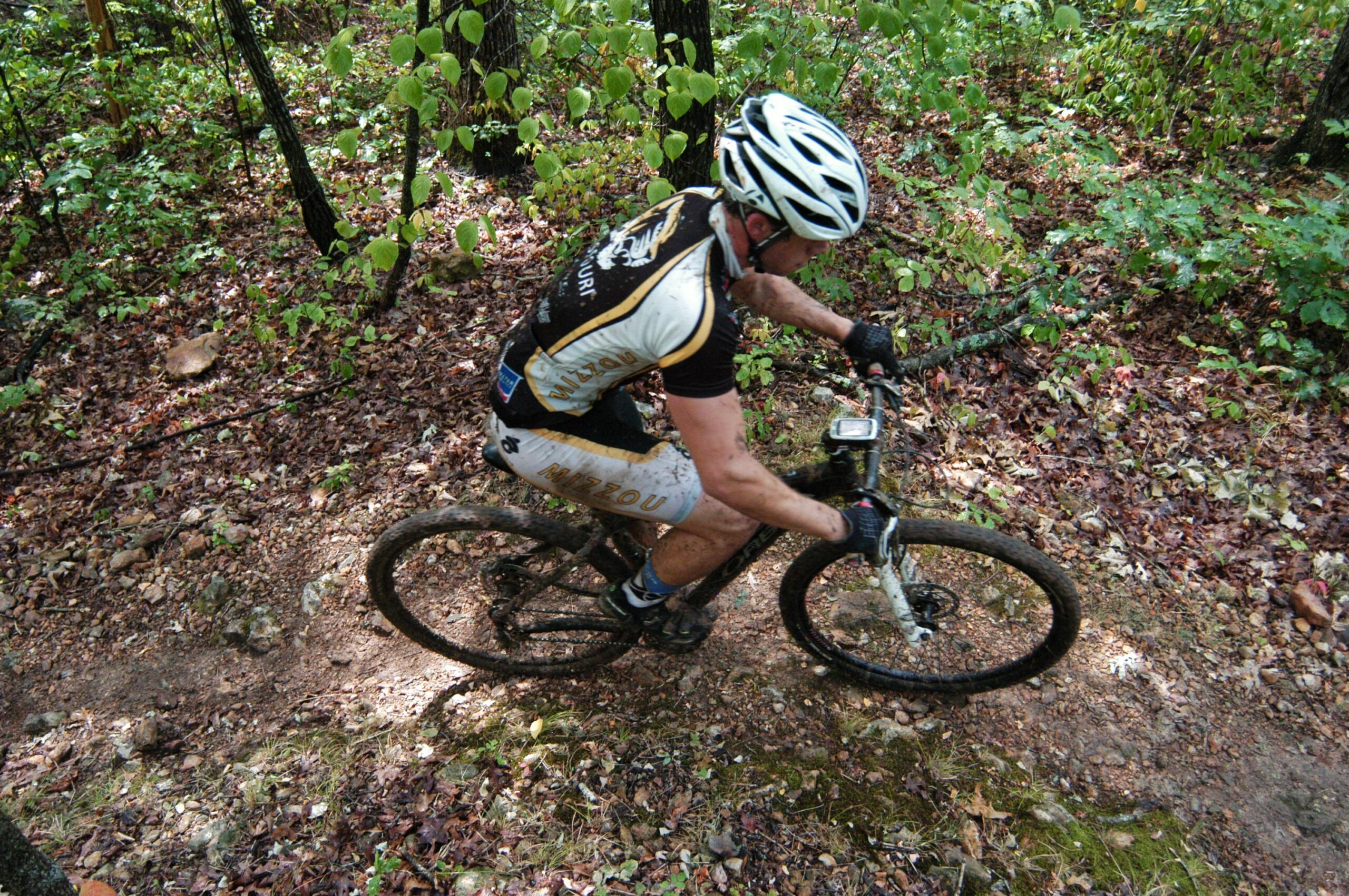 A mountain biker riding on a narrow, dirt trail surrounded by greenery and fallen leaves. The cyclist is wearing a black and white jersey with "Mizzou" printed on it, and is covered in mud from riding. The image is captured from an overhead angle, showcasing the bike's details and the rugged terrain. Binder Lake mountain bike trail.