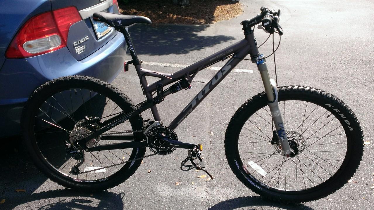 Titus Moto Lite: Mountain bike parked next to a blue car in a parking lot. The bike features a gray frame, thick tires designed for off-road use, and a visible suspension system. The background includes pavement and some vegetation.