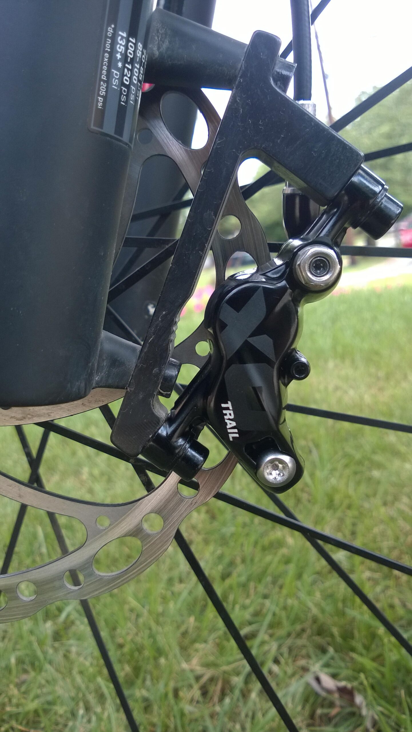 Specialized Rockhopper Pro EVO 29: Close-up view of a bicycle brake caliper mounted on a disc brake, showing the intricate details of the black brake component labeled "TRAIL" alongside a metallic disc with holes, set against a grassy background.