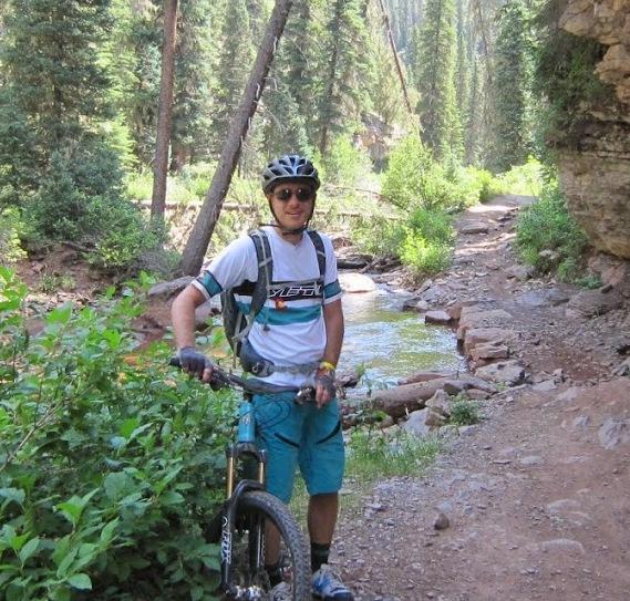 Yeti SB66: A person wearing a helmet and sunglasses stands next to a mountain bike on a rocky trail surrounded by lush greenery and tall trees. A small stream runs alongside the path, and sunlight filters through the trees in the background.