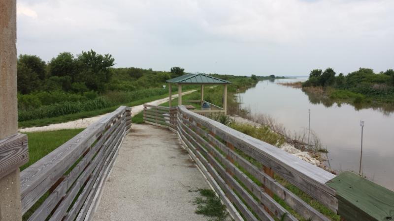 A serene pathway leading to a gazebo beside a calm river, surrounded by lush greenery and gentle slopes. The walkway, bordered by wooden railings, guides the viewer toward the water's edge, under a cloudy sky. Lake Apopka Restoration Area mountain bike trail.