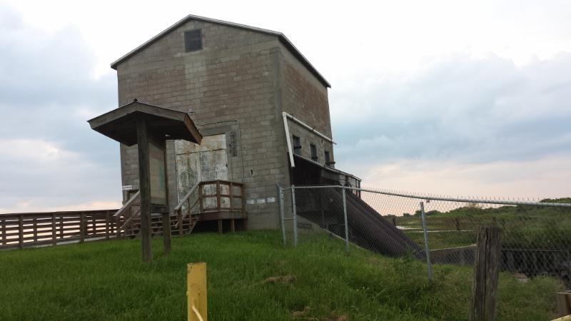 Alt text: A large, grey concrete structure with a sloped roof, standing on a grassy hill. A wooden deck with stairs leads to a door at the front, and a metal fence encloses the area. The sky is overcast with clouds. Lake Apopka Restoration Area mountain bike trail.