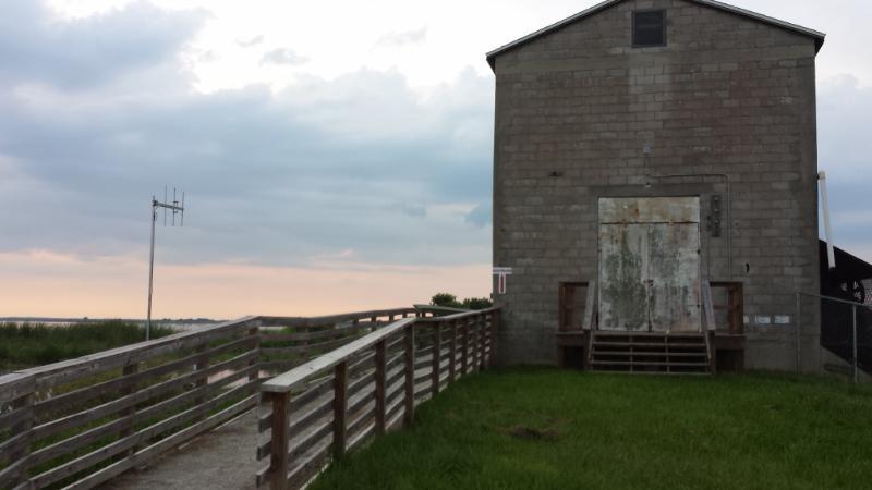 A weathered building made of gray stone, featuring a large, rusty door, is set against a cloudy sky. A wooden walkway with railings leads up to the building, surrounded by green grass and a glimpse of water in the distance. Lake Apopka Restoration Area mountain bike trail.
