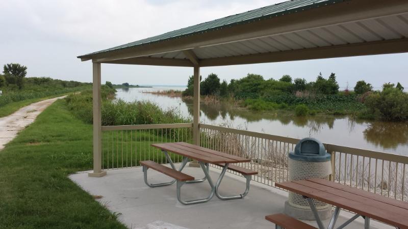 A riverside pavilion with a green roof and picnic tables overlooks a calm body of water surrounded by lush vegetation. A walking path runs along the water's edge, leading into a natural landscape. Lake Apopka Restoration Area mountain bike trail.