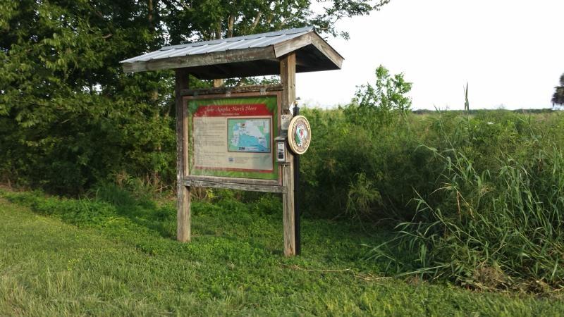 A wooden information sign located beside a grassy area, featuring a map and details about the nearby area. The sign is partially shaded by a slanted roof, surrounded by trees and tall grass. Lake Apopka Restoration Area mountain bike trail.