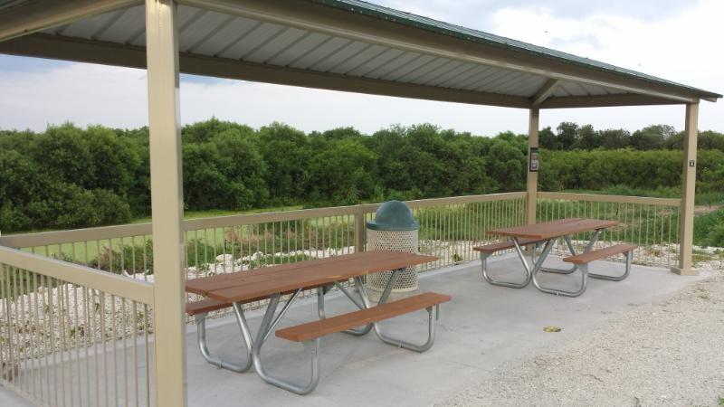 A covered picnic area featuring two wooden picnic tables with metal frames, situated on a concrete surface. In the background, there is a lush green landscape with trees. A trash can is placed to the side of the picnic area. The sky is partly cloudy, creating a serene outdoor setting. Lake Apopka Restoration Area mountain bike trail.