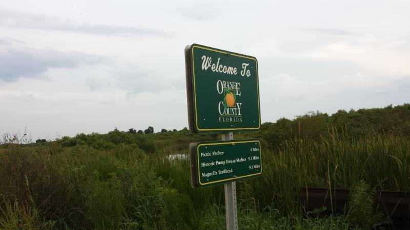 A green welcome sign for Orange County, Florida, featuring an orange graphic. The sign includes directions to nearby attractions such as the picnic shelter, historic pump house, and Magnolia Trailhead, with distances noted. The background features a natural landscape with tall grass and a cloudy sky. Lake Apopka Restoration Area mountain bike trail.