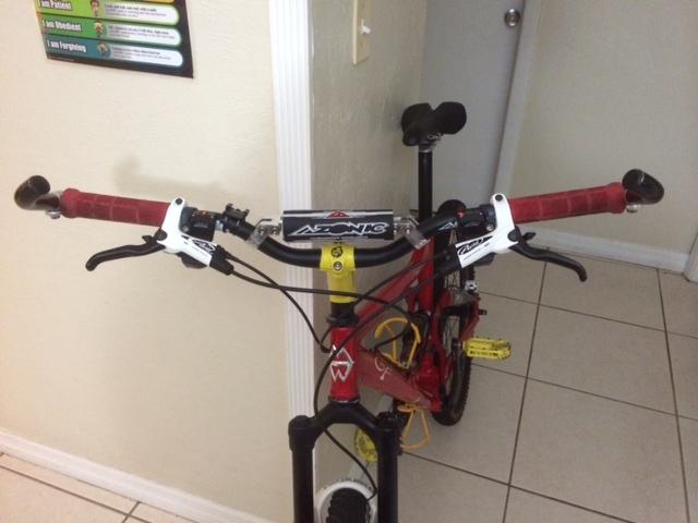Alt tag: "Close-up view of the handlebars of a red mountain bike with black grips, brake levers, and a yellow and black top tube sticker, set against a plain wall with a door in the background."