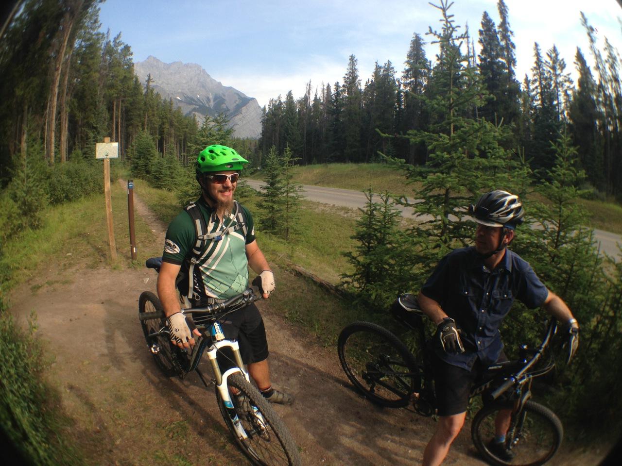 Two mountain bikers are standing on a dirt trail surrounded by tall trees. One biker, wearing a bright green helmet and a patterned shirt, is smiling and holding his bike. The other biker is in a dark blue shirt and black shorts, leaning against his bike. A signpost is visible in the background, with a mountain range visible in the distance under a partly cloudy sky. Tunnel Mountain Trail System mountain bike trail.