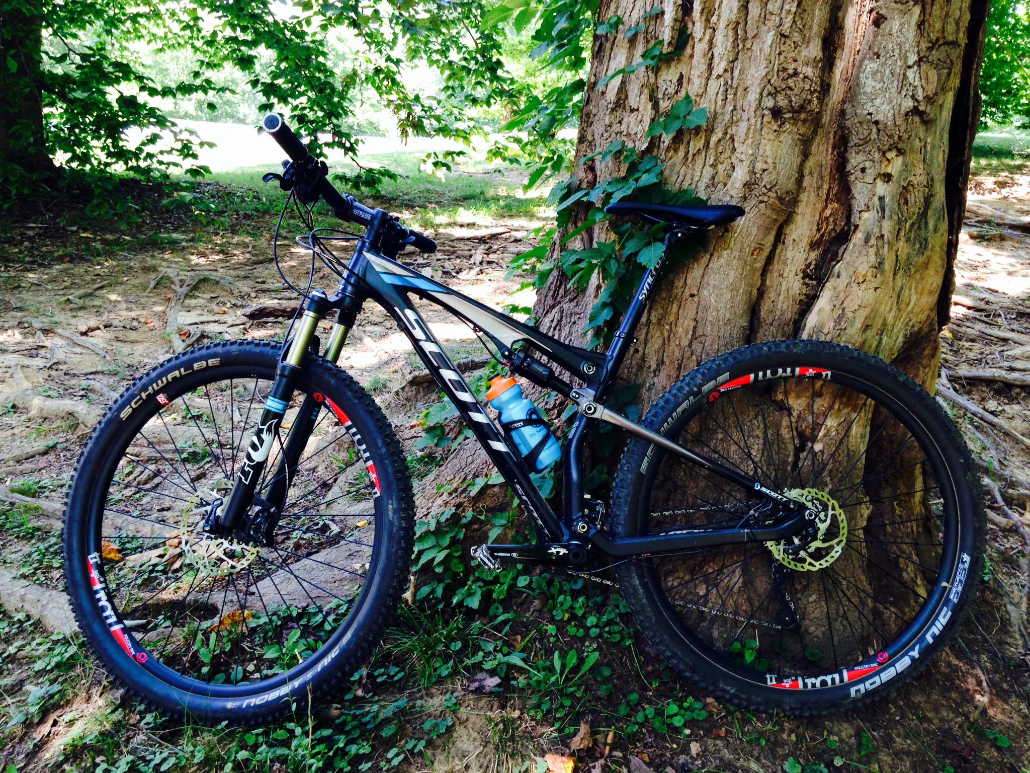 A black mountain bike leaning against a large tree, surrounded by green foliage. The bike features thick tires designed for off-road terrain, a visible water bottle, and sunlight filtering through the trees in the background. Cherokee Park mountain bike trail.