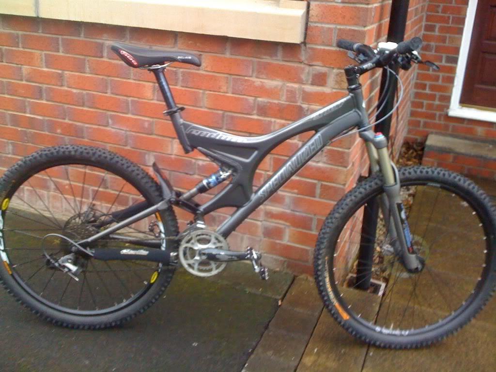 Specialized Enduro SL Comp FSR: A modern mountain bike in grey and black, leaning against a brick wall. The bike features a prominent suspension system, thick tires suitable for off-road terrain, and a sleek frame design. The background includes a wooden path and a door, suggesting a residential setting.