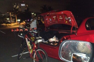 Giant Boulder SE: A cyclist stands beside a red car with its trunk open, showcasing various stickers on the trunk. A bicycle with a helmet and other gear is parked nearby. The scene is illuminated by ambient city lights, indicating it is nighttime.