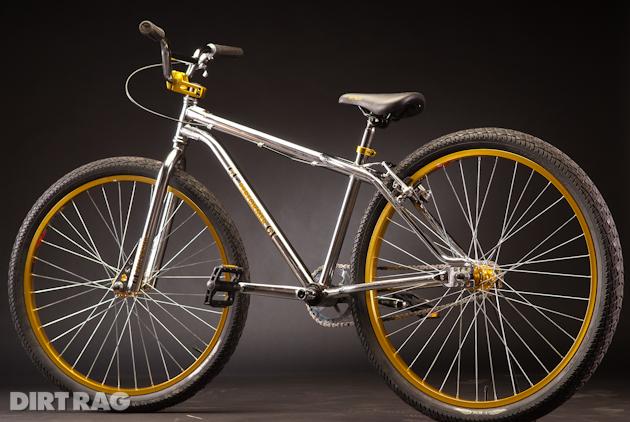 GT Performer 26: A shiny silver bicycle with yellow accents, featuring large wheels and a simple design. The bike is positioned against a dark background, showcasing its sleek frame and distinctive tires. The handlebars are equipped with grips, and the seat is slightly elevated for a comfortable riding position.