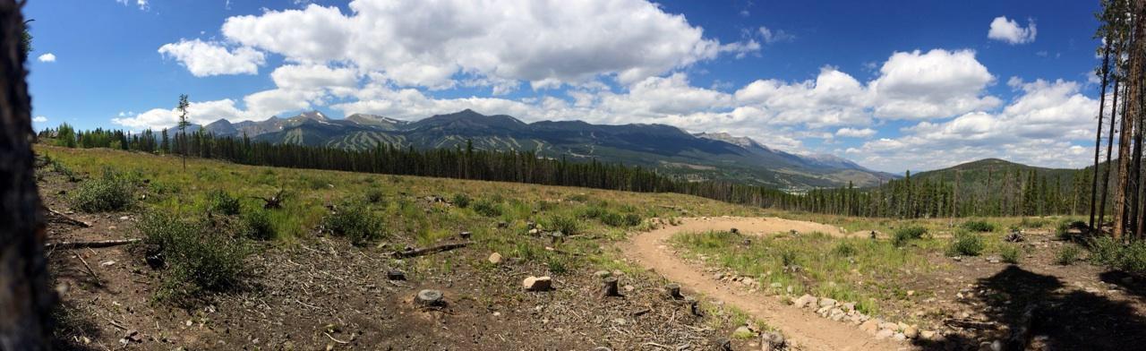 A panoramic view of a mountainous landscape under a blue sky, featuring rolling hills, patches of green vegetation, and a winding dirt path. Fluffy white clouds are scattered across the sky, and the scene conveys a serene and natural outdoor setting. Barney Ford / Juniata Trail mountain bike trail.