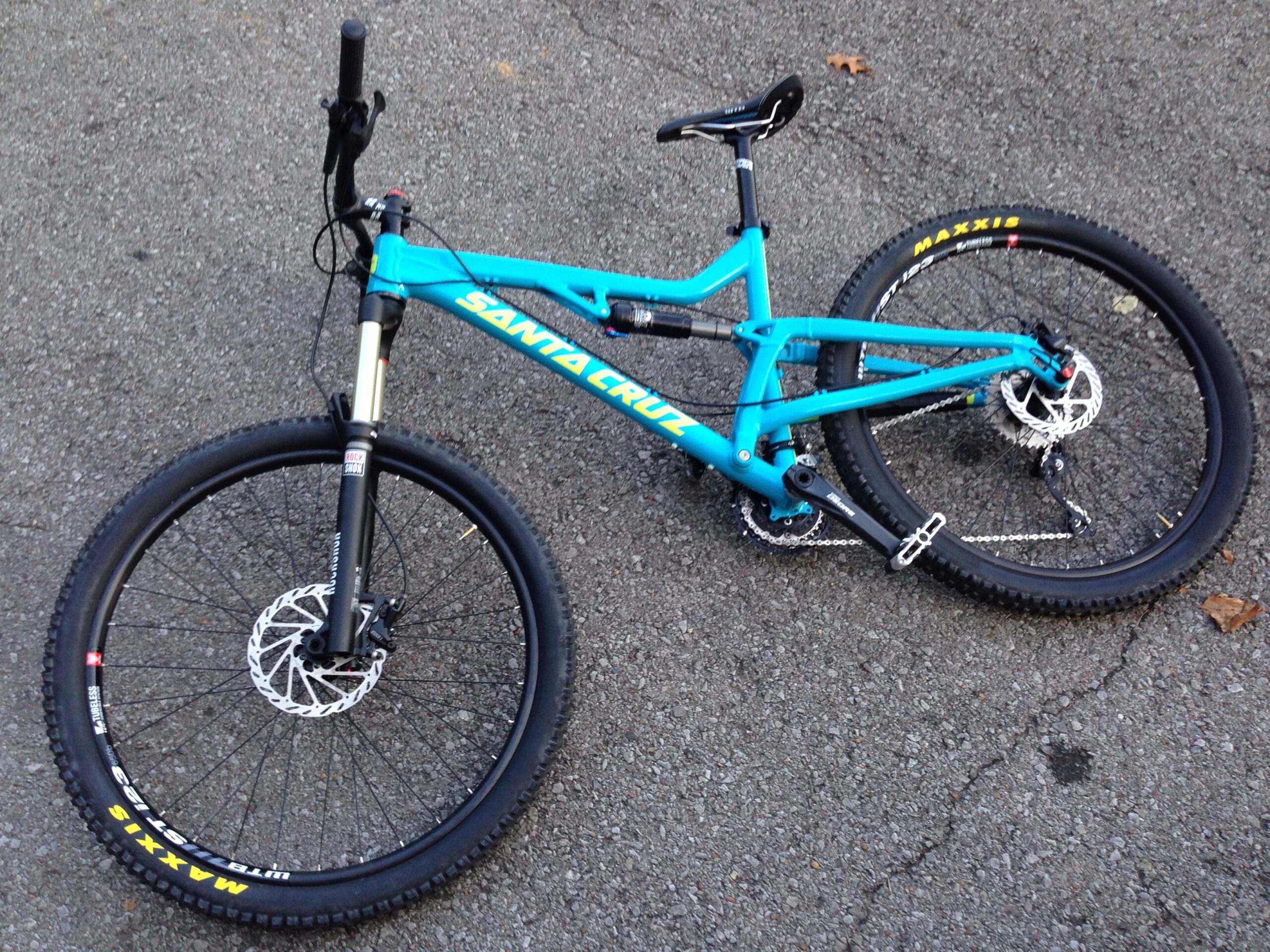 Santa Cruz Heckler: A bright blue Santa Cruz mountain bike lying on a gravel surface, featuring thick black Maxxis tires and a suspension fork. The bike's frame showcases a sleek design, and the handlebars are equipped with gear shifters and brake levers.