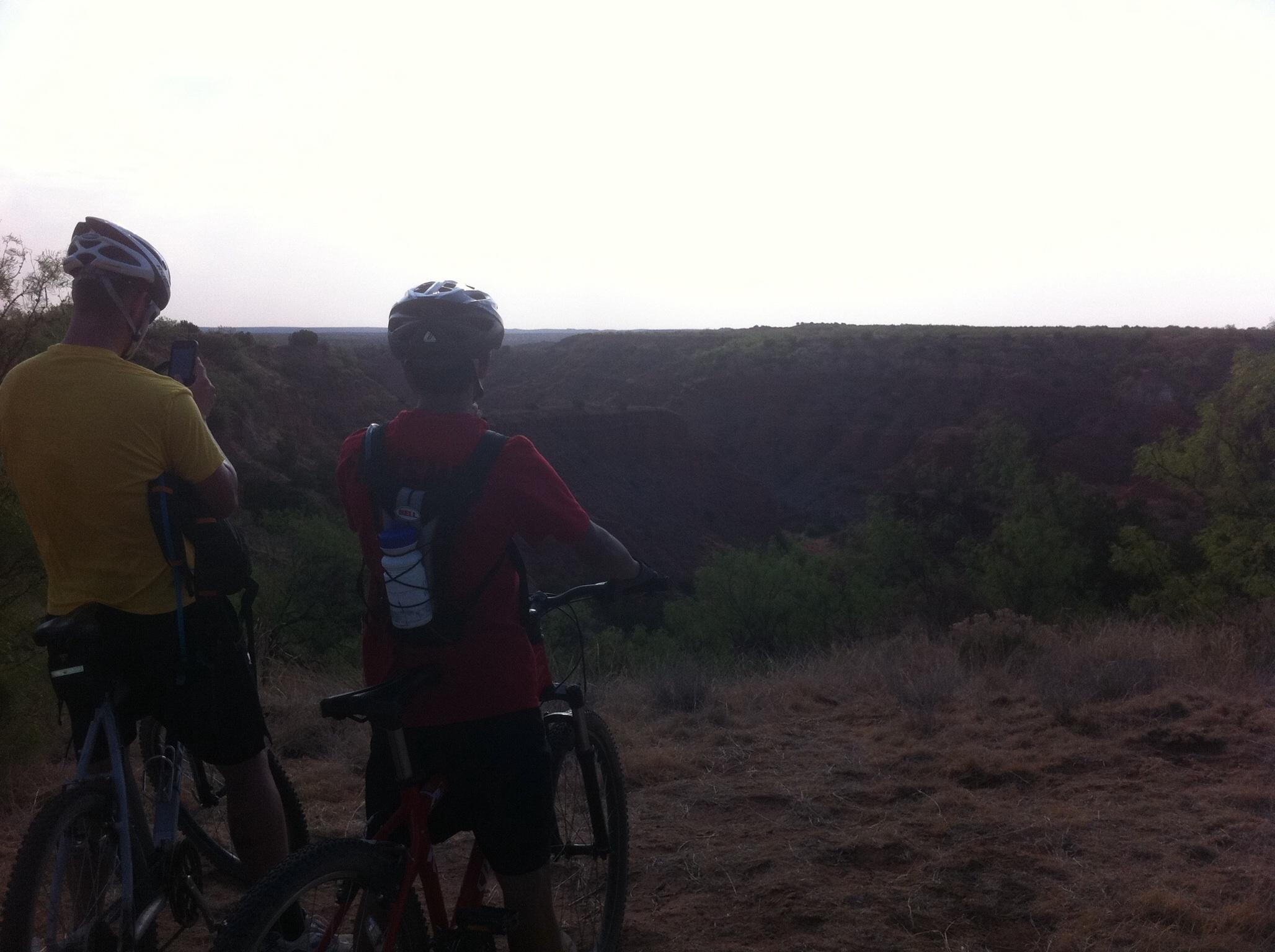 Two mountain bikers, one in a yellow shirt and the other in a red shirt, stand on a dirt hillside overlooking a canyon. The biker on the left is using a smartphone to take a photo, while the other is gazing at the scenery. The landscape features rugged terrain with green bushes and a hazy sky in the background. Palo Duro Canyon mountain bike trail.