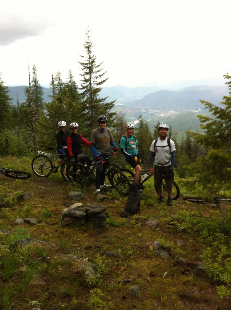 A group of five mountain bikers posing for a photo on a forested trail, with their bikes resting nearby. A dog is sitting in front of them, and the background features rolling hills and greenery under a cloudy sky. Swine Flu Loop mountain bike trail.