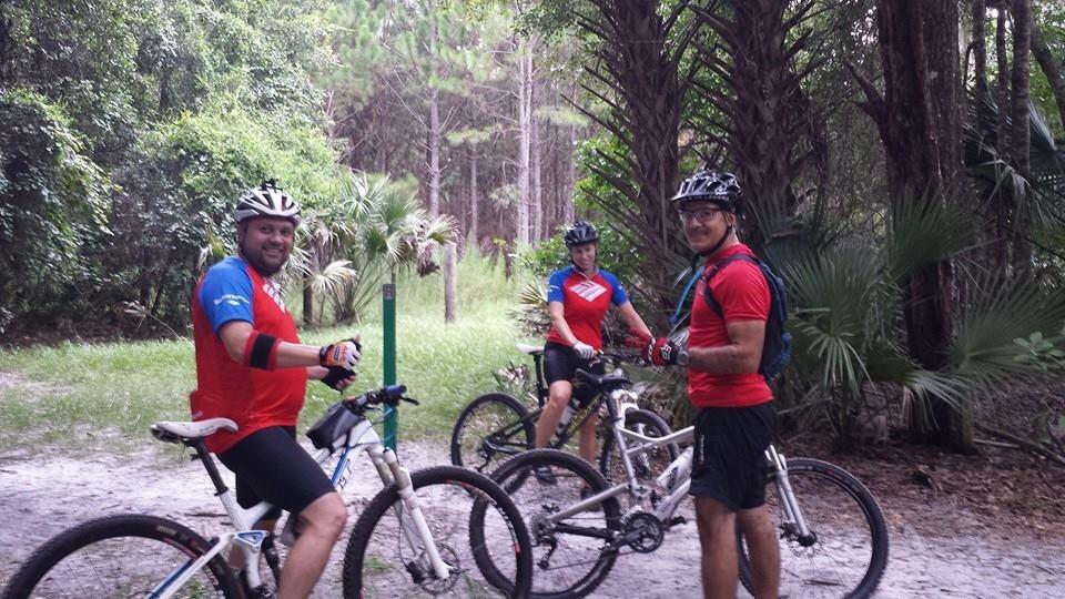 Three mountain bikers in red and blue jerseys pose with their bicycles in a wooded area. They are smiling and standing on a dirt path surrounded by trees and lush greenery. The background includes palm and pine trees, indicating a natural outdoor setting. Little Big Econ State Forest mountain bike trail.
