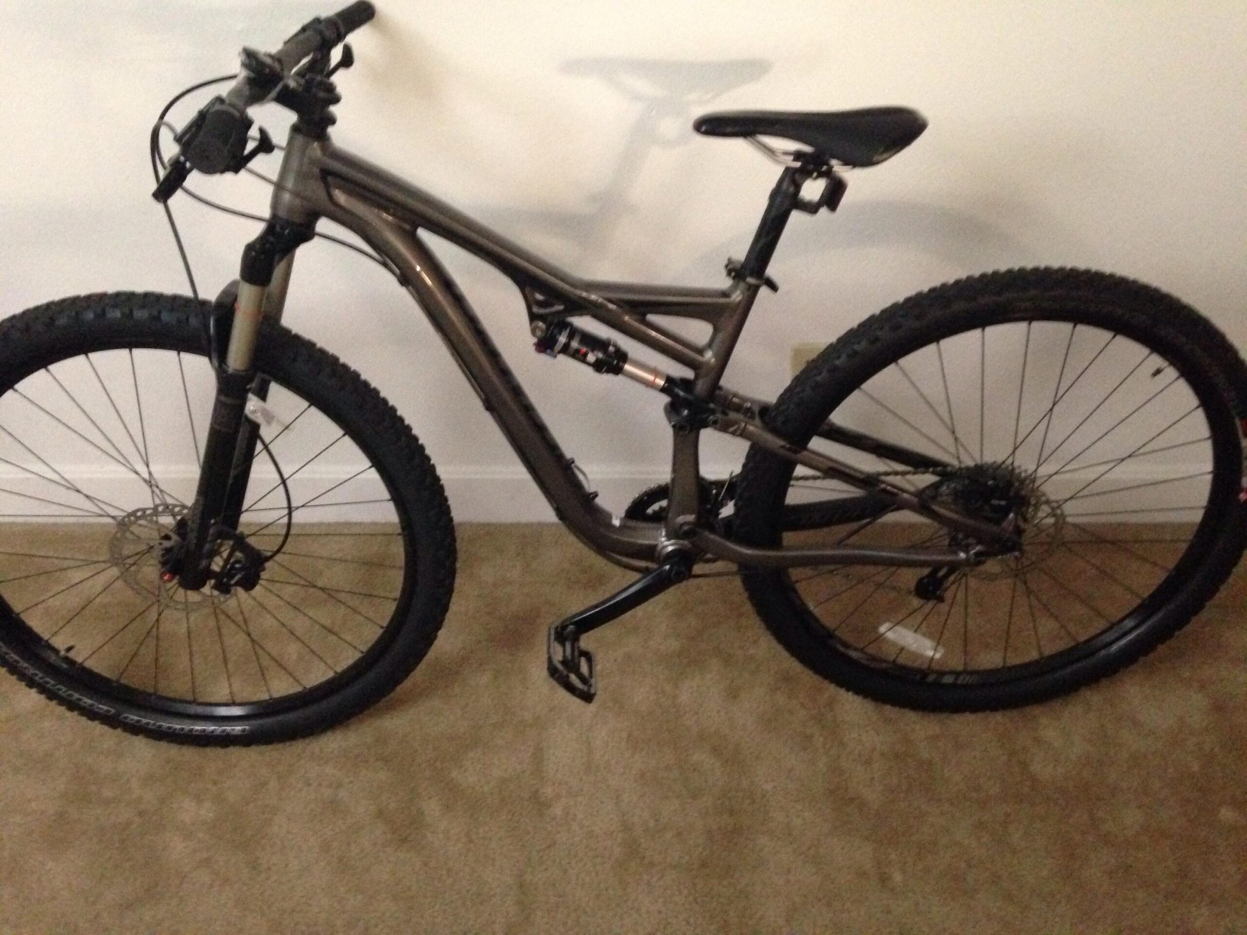 Specialized Camber Comp 29: A modern mountain bike with a sleek, metallic gray frame, featuring front suspension forks and large, knobby tires. The bike is positioned on a beige carpeted floor against a plain white wall, showcasing its design and components.
