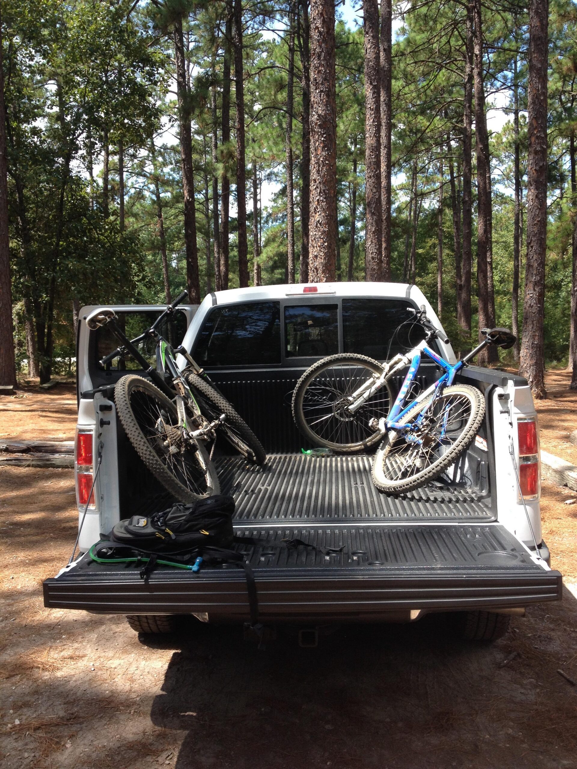 A white pickup truck parked in a wooded area, with two mountain bikes secured in the bed of the truck. A black backpack is placed on the truck's tailgate, surrounded by trees and a clear sky in the background. Harbison State Forest mountain bike trail.