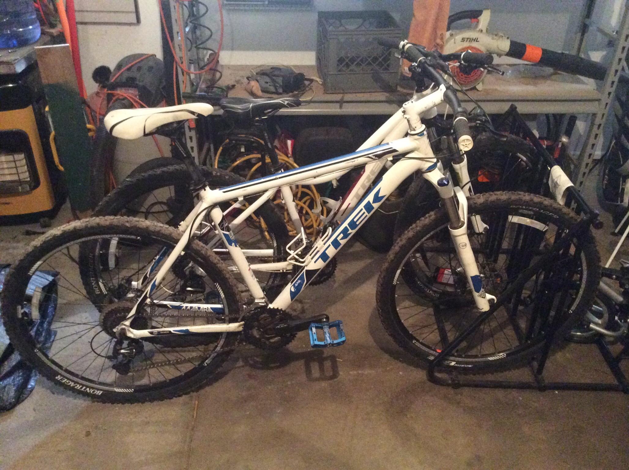 Trek Mamba: A white Trek mountain bike with blue accents, leaning against another bike, in a cluttered garage. The background shows various tools and equipment, including a heater and some electrical cords.
