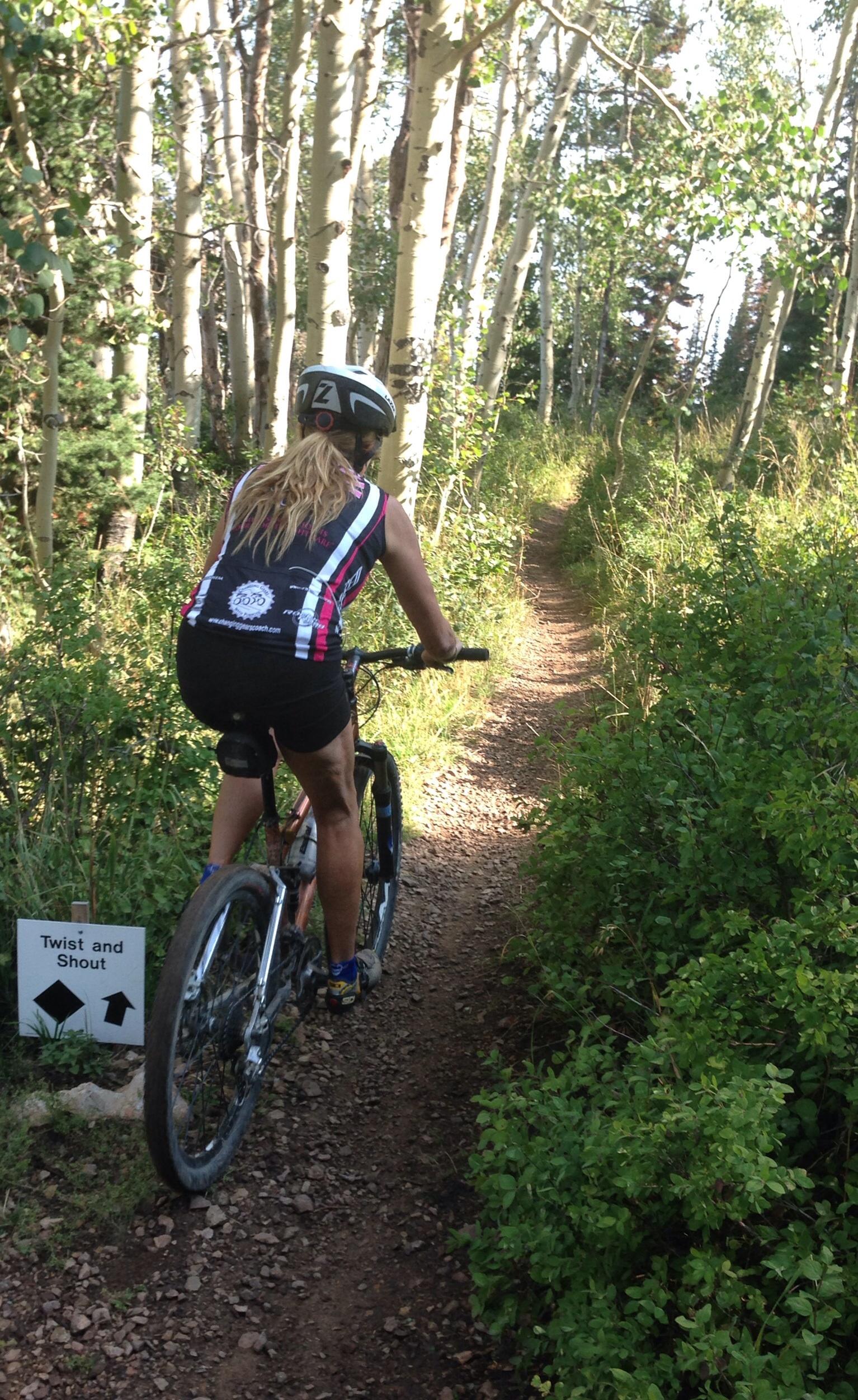 A person on a mountain bike rides along a narrow dirt trail surrounded by lush greenery and tall aspen trees. A sign labeled "Twist and Shout" is positioned beside the trail, indicating the path's name. The cyclist wears a black helmet and a sleeveless jersey with colorful stripes. Deer Valley Resort Bike Park mountain bike trail.