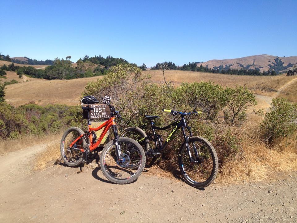 Two mountain bikes, one orange and one black, are parked next to a sign that reads "Bicycles Must Stay on Trail" amidst a scenic landscape of rolling hills and greenery. The background features a clear blue sky and dry grassy fields. Camp Tamarancho mountain bike trail.
