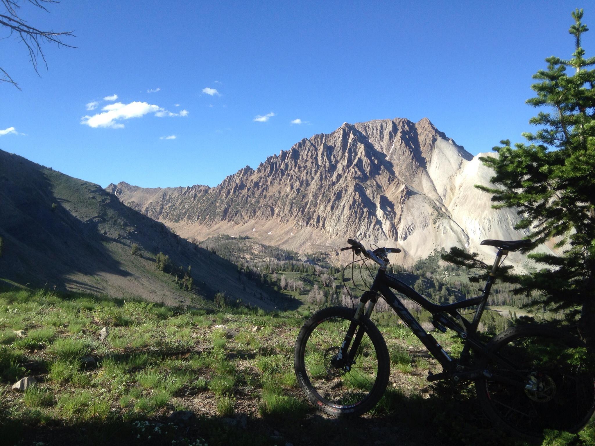 Yeti 575: A mountain bike is parked on a grassy area with a backdrop of rugged mountain peaks under a clear blue sky with a few scattered clouds. The scene showcases the beauty of nature, with lush greenery in the foreground and steep, rocky slopes in the background.