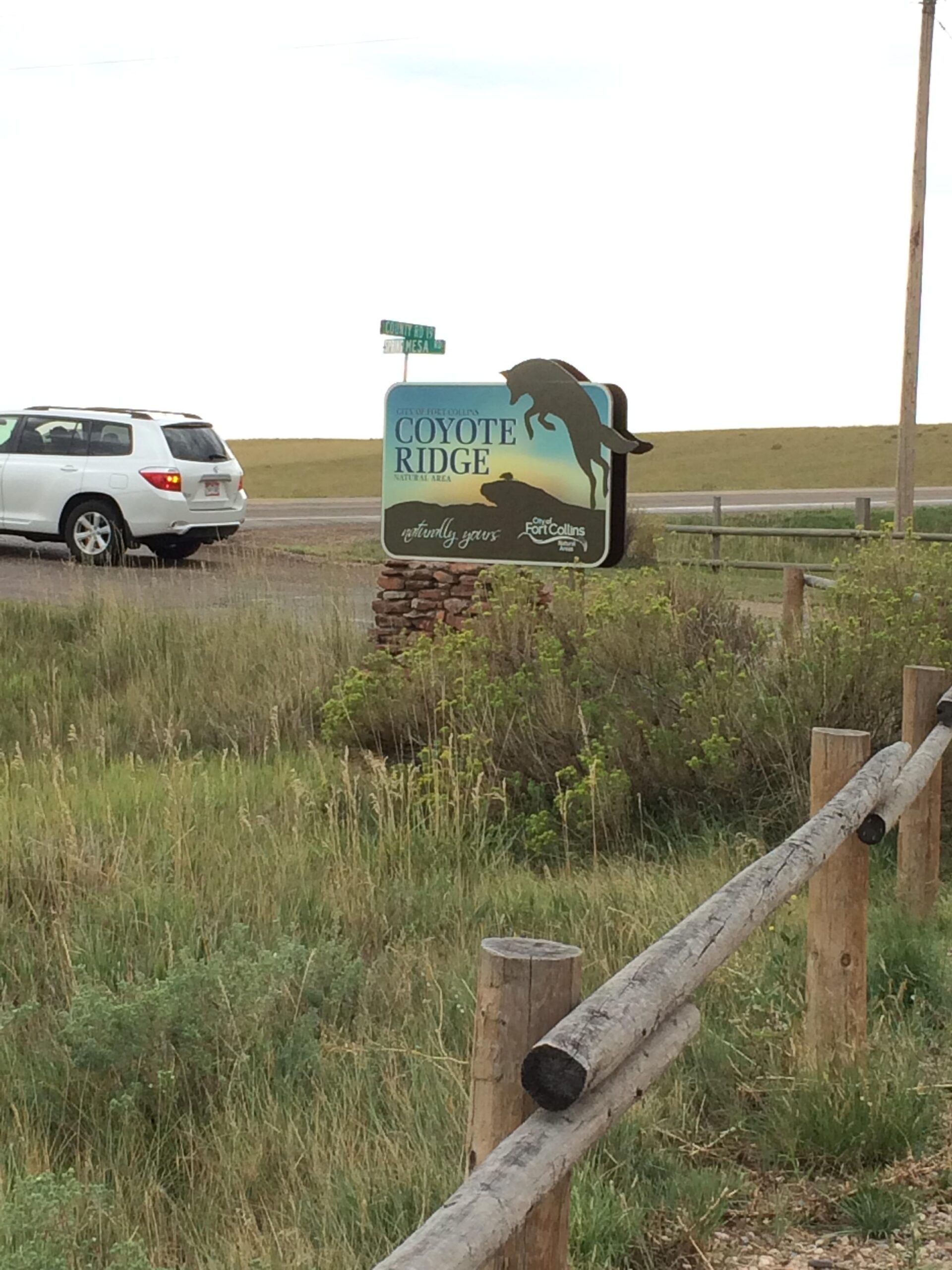 Sign for Coyote Ridge Natural Area, featuring a silhouette of a coyote jumping, alongside a road with a white SUV parked nearby. The background shows grassy fields and a wooden fence. Coyote Ridge mountain bike trail.