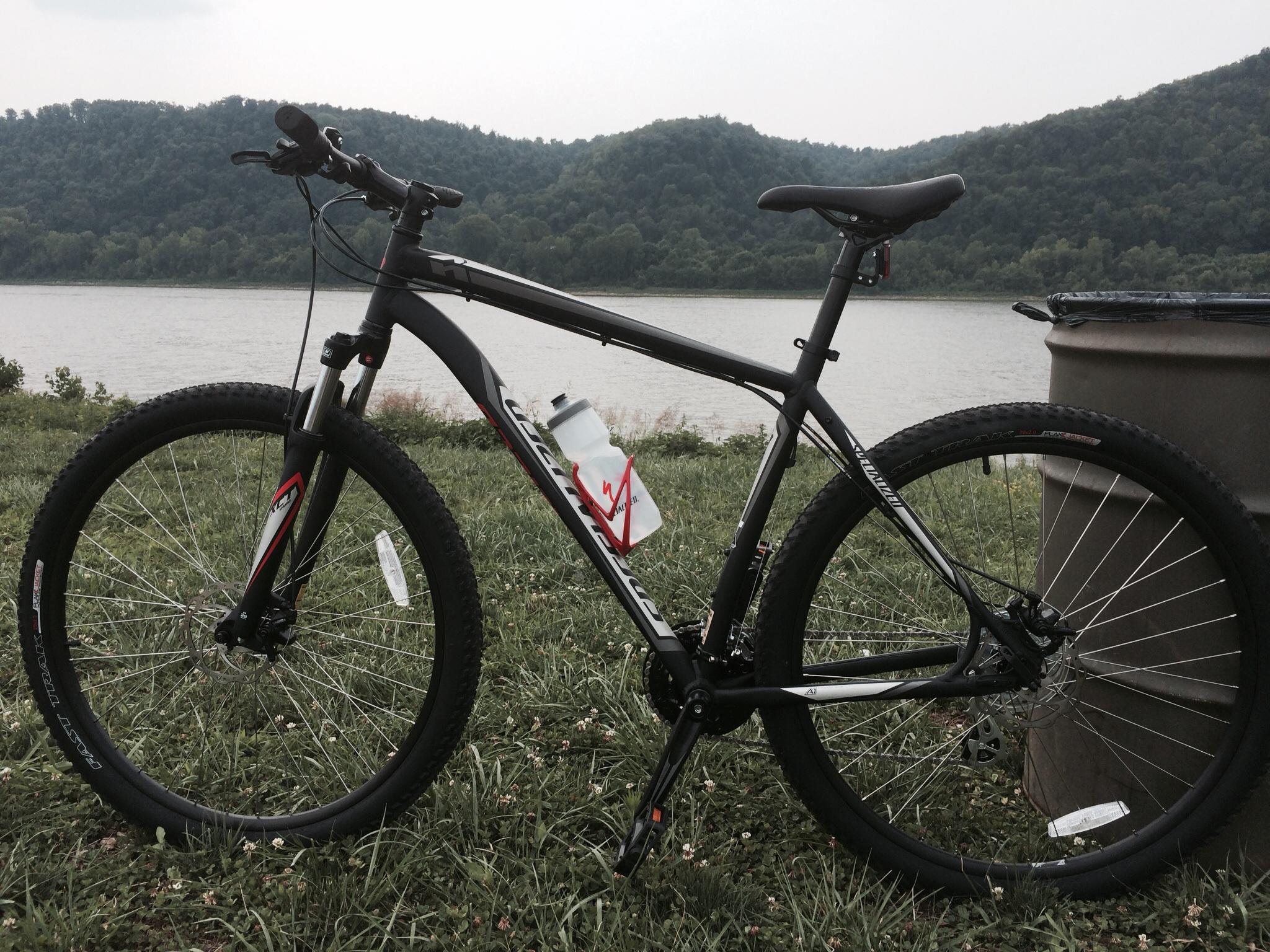 Specialized Hardrock Sport Disc 29er: A black mountain bike is positioned on grassy ground near a riverbank, with hills in the background. A water bottle is attached to the bike frame, and a trash can is visible nearby. The scene is set on a cloudy day.