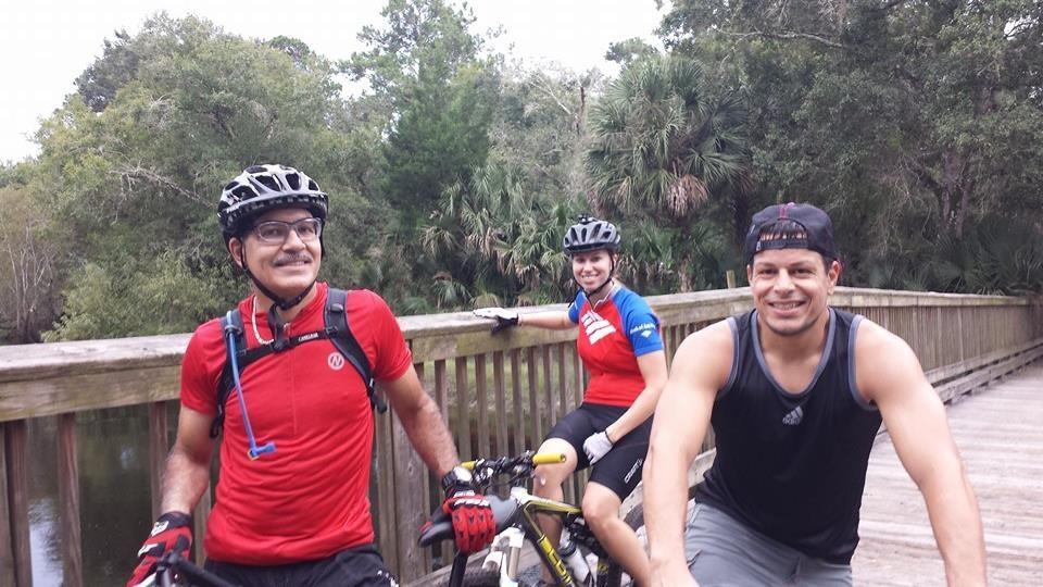 Three friends posing on a wooden bridge during a mountain biking trip in a lush green area. They are wearing cycling gear and helmets, with two men in red and black shirts and one woman in a blue and red shirt. Trees and foliage are visible in the background, creating a natural setting. Little Big Econ State Forest mountain bike trail.