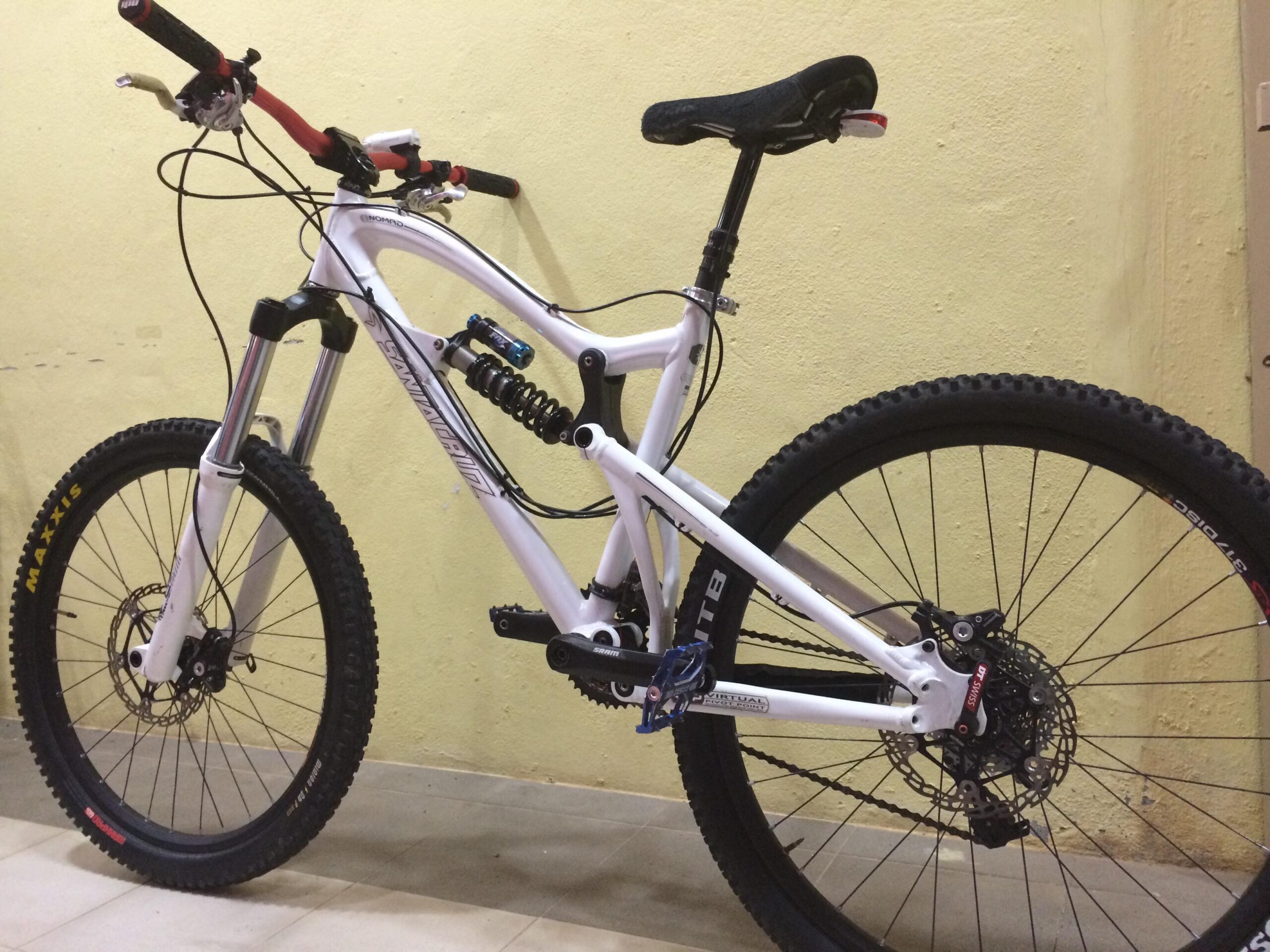 Santa Cruz Nomad: A white mountain bike with a dual suspension frame, equipped with Maxxis tires and disc brakes. The bike features a sleek design with red accents on the handlebars and seat. It is positioned against a light yellow wall, showcasing its features and components.