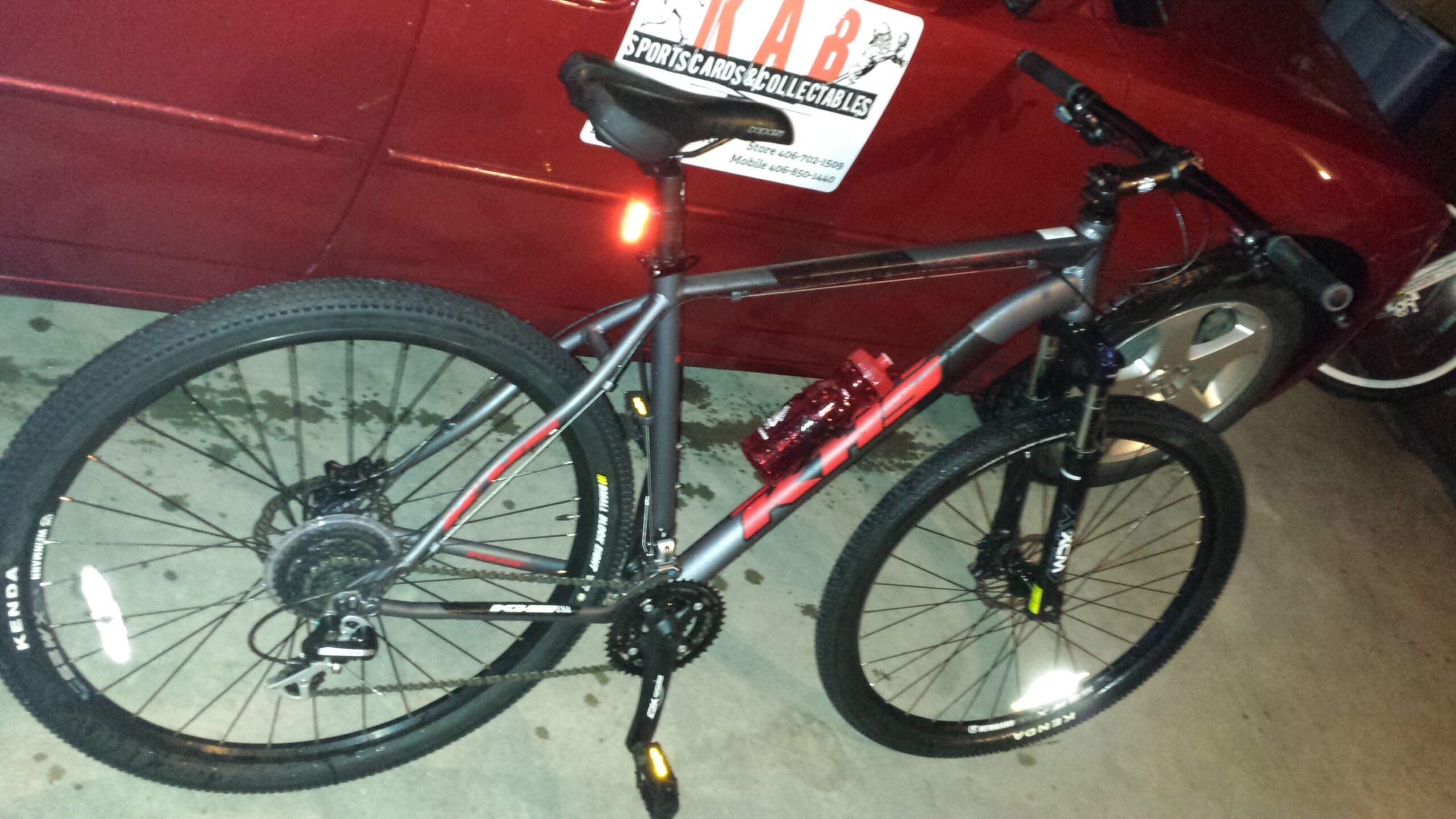 KHS Winslow: Mountain bike parked beside a red car in a garage. The bike features a gray and red frame, black tires, and a water bottle attached to the side. The garage floor has some water stains, and a sign with "KAB Sports Cards & Collectables" is visible in the background.