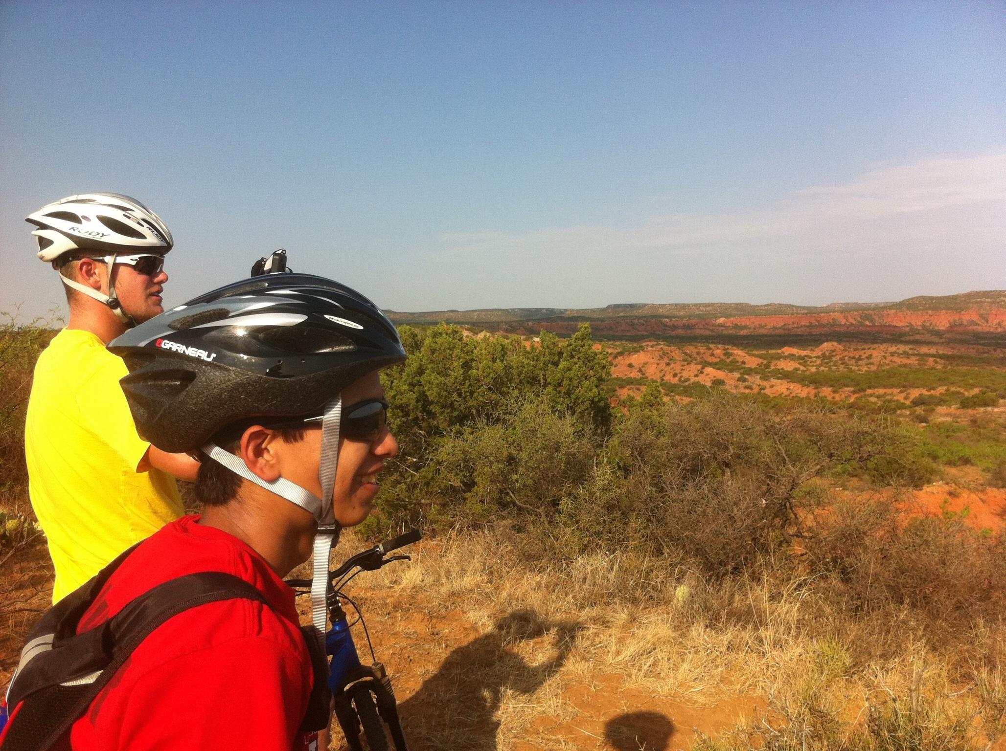 Two mountain bikers wearing helmets and sunglasses stand on a ridge overlooking a scenic view of red rock formations and greenery. One biker is in a yellow shirt and the other in a red shirt, both gazing into the distance. The sky is clear with a few clouds, and the landscape features dry grass and shrubs typical of a desert-like environment. Palo Duro Canyon mountain bike trail.