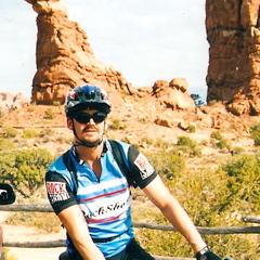 Specialized Stumpjumper Comp: A cyclist wearing a blue and black jersey and a helmet poses on a trail, with a large rock formation in the background. The scene captures a sunny day in a natural setting, surrounded by desert vegetation.