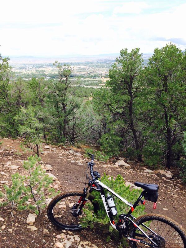 A mountain bike resting next to a gravel path, surrounded by tall green trees, with a scenic view of valleys and mountains in the background under a cloudy sky. Ojitos Canyon mountain bike trail.