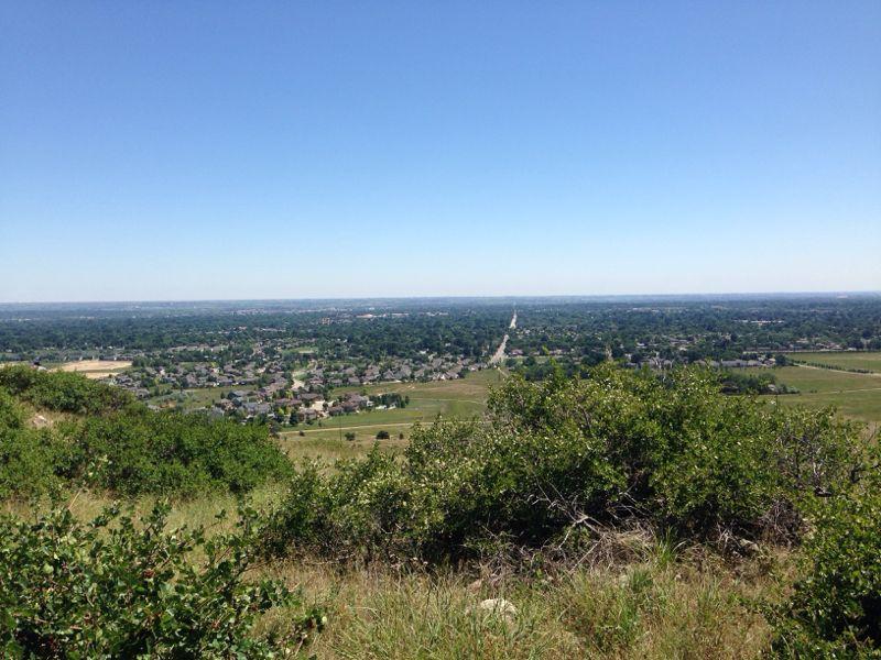 A panoramic view from a hilltop, showcasing a vast landscape dotted with residential neighborhoods, fields, and roads under a clear blue sky. The lush greenery in the foreground contrasts with the urban expanse in the distance. Maxwell's mountain bike trail.
