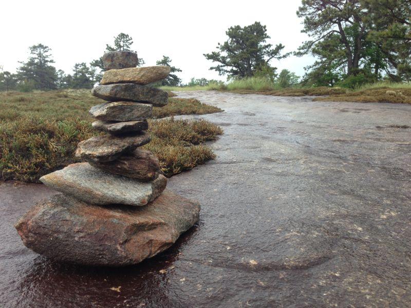 A carefully stacked pile of stones sits on a flat, wet surface, surrounded by sparse vegetation and trees in the background. The scene is overcast, suggesting a calm, natural outdoor setting. DuPont State Recreational Forest mountain bike trail.