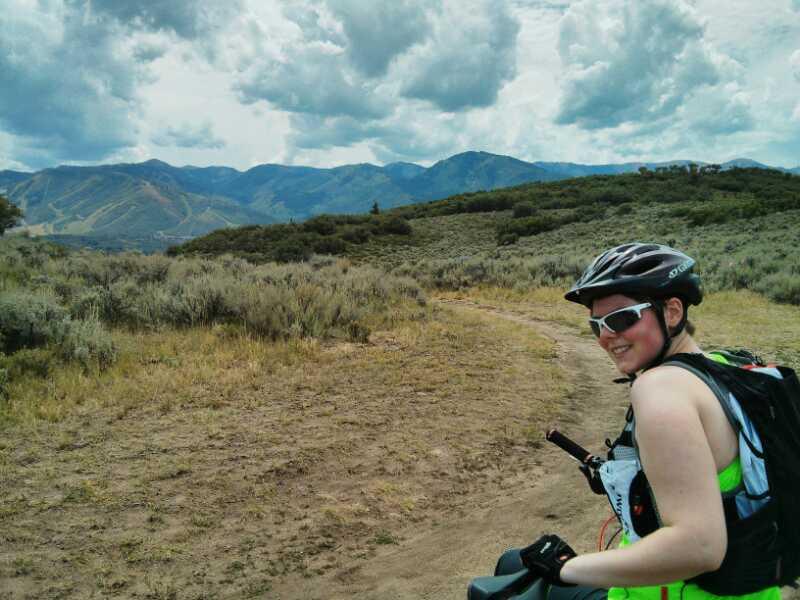 A person in a cycling helmet and sunglasses smiles while standing beside a mountain biking path. The background features rolling hills and a cloudy sky, with shrubs and grass surrounding the trail. Round Valley mountain bike trail.