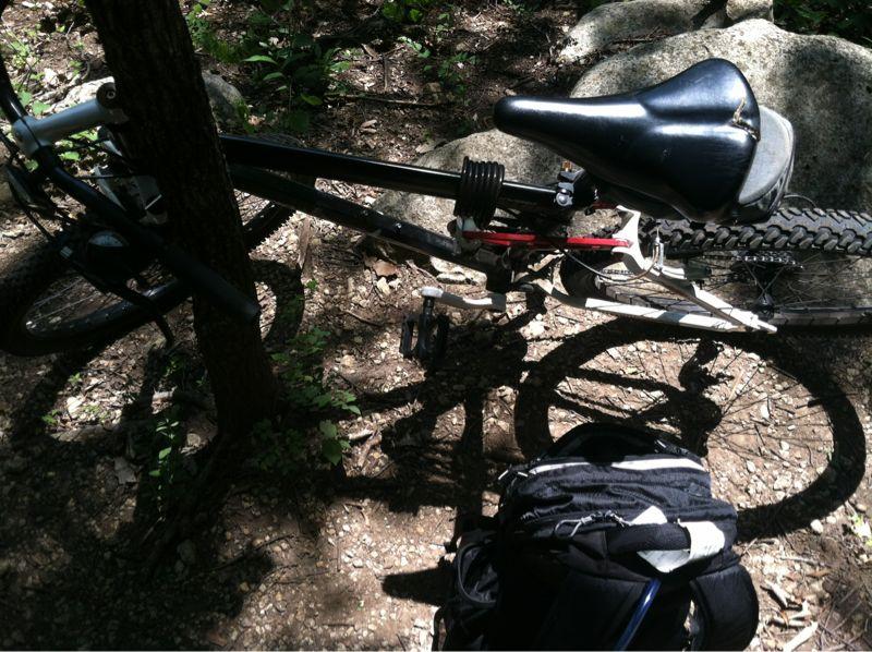 A mountain bike leaning against a tree on a dirt path, surrounded by rocks and greenery. A black and white backpack is positioned on the ground nearby, in dappled sunlight. Shadows cast by the tree and the bike create an intriguing play of light. Camp Horizon mountain bike trail.