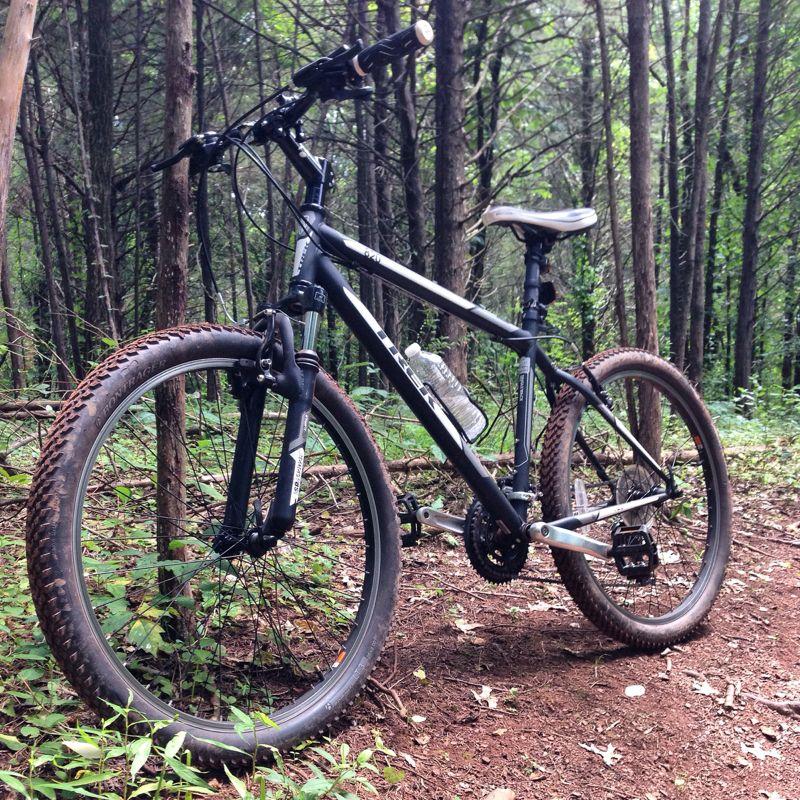 A mountain bike parked on a dirt trail surrounded by tall trees and greenery. A water bottle is attached to the frame, indicating it’s ready for outdoor adventures. The bike's tires are designed for rugged terrain. Six Mile Run mountain bike trail.