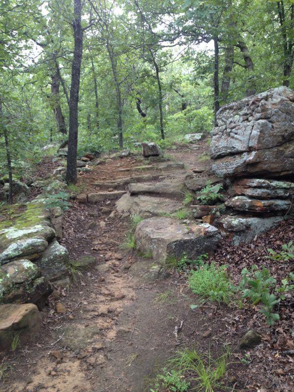 A winding trail through a lush forest, featuring rocky steps and large boulders on either side. The path is surrounded by green foliage and trees, with a natural earthy surface. Osage Hills State Park mountain bike trail.
