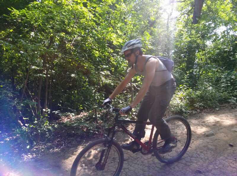 A person riding a mountain bike on a dirt trail surrounded by lush greenery. The cyclist is wearing a helmet and a sleeveless shirt, with a backpack visible on their back. Sunlight filters through the trees, creating a bright and vibrant atmosphere. Palos Forest Preserve mountain bike trail.