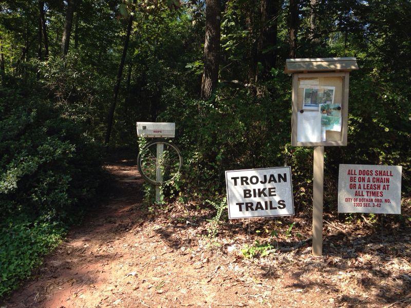 Entrance to the Trojan Bike Trails, featuring a sign indicating the trail name, an information board with postings, and a notice about dog leash regulations. Surrounded by dense greenery and trees, the path leads into a natural setting. Troy State University Dothan Trails mountain bike trail.