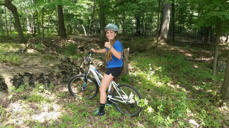 A young person wearing a helmet gives a thumbs-up while sitting on a mountain bike in a lush green forest setting. Sunlight filters through the trees, creating a vibrant outdoor atmosphere. Chimney Rock mountain bike trail.