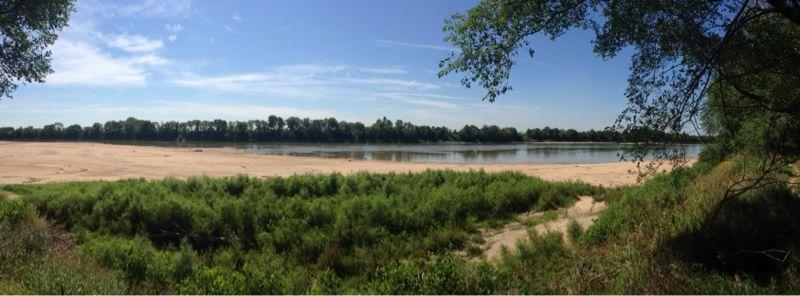 A panoramic view of a serene river landscape with sandy banks and lush green vegetation along the shore. The scene features clear blue skies with a few wispy clouds, reflecting a peaceful day by the water. Lawrence Riverfront Trails mountain bike trail.